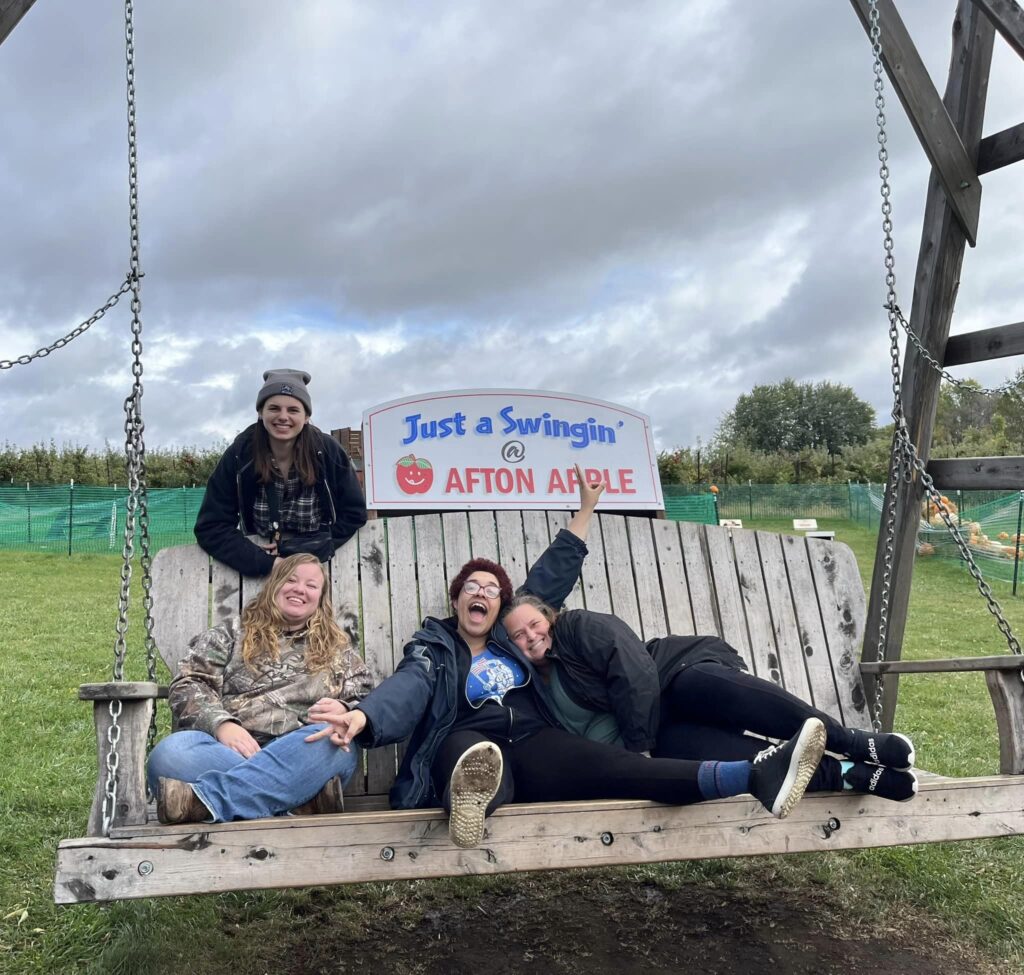 four women smiling, three sitting on a bench and one leaning over the back of it. Sign on the bench reads "Just a Swingin' Afton Apple"